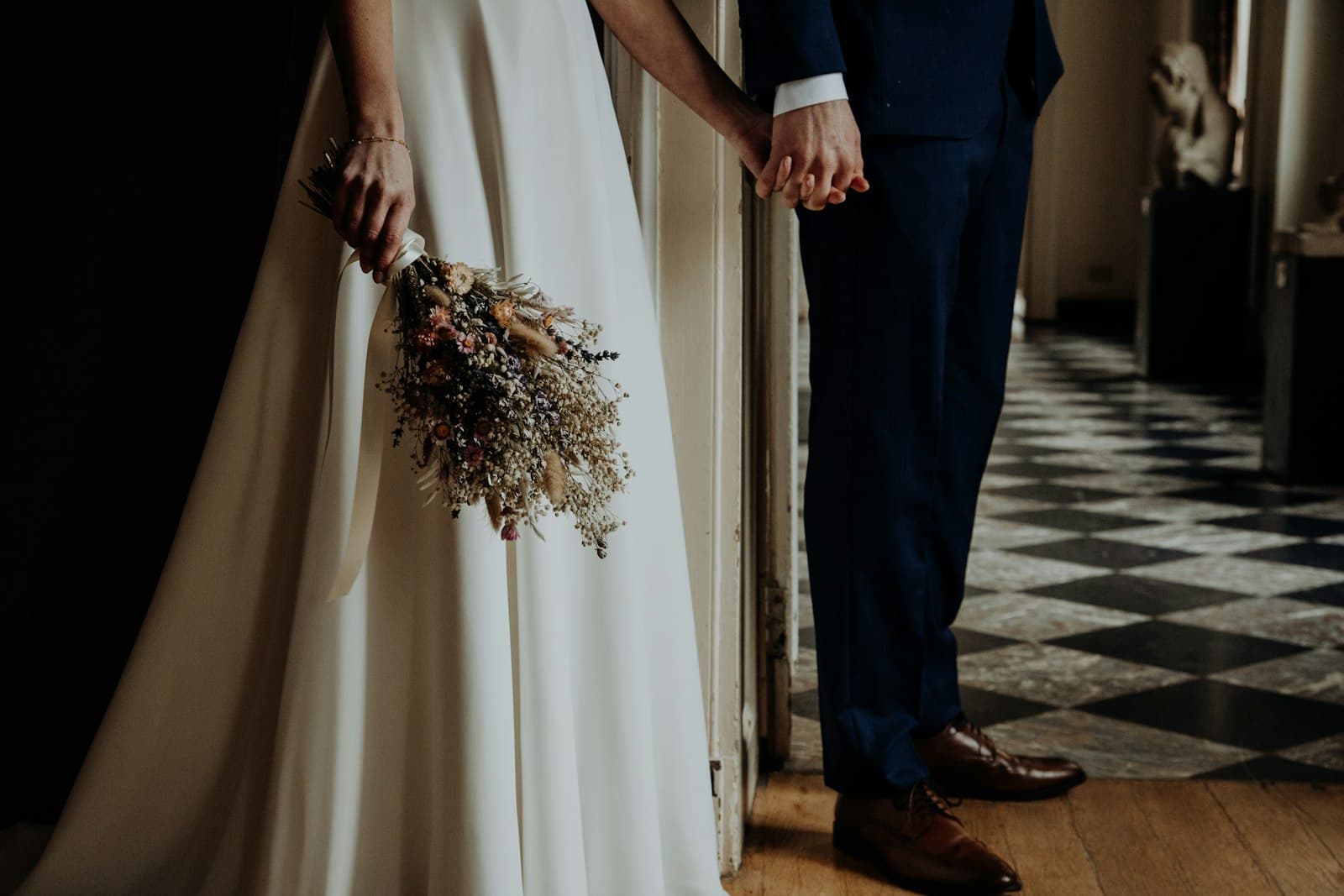 Mariée en robe blanche tenant un bouquet de fleurs séchées, main dans la main avec le marié en costume bleu