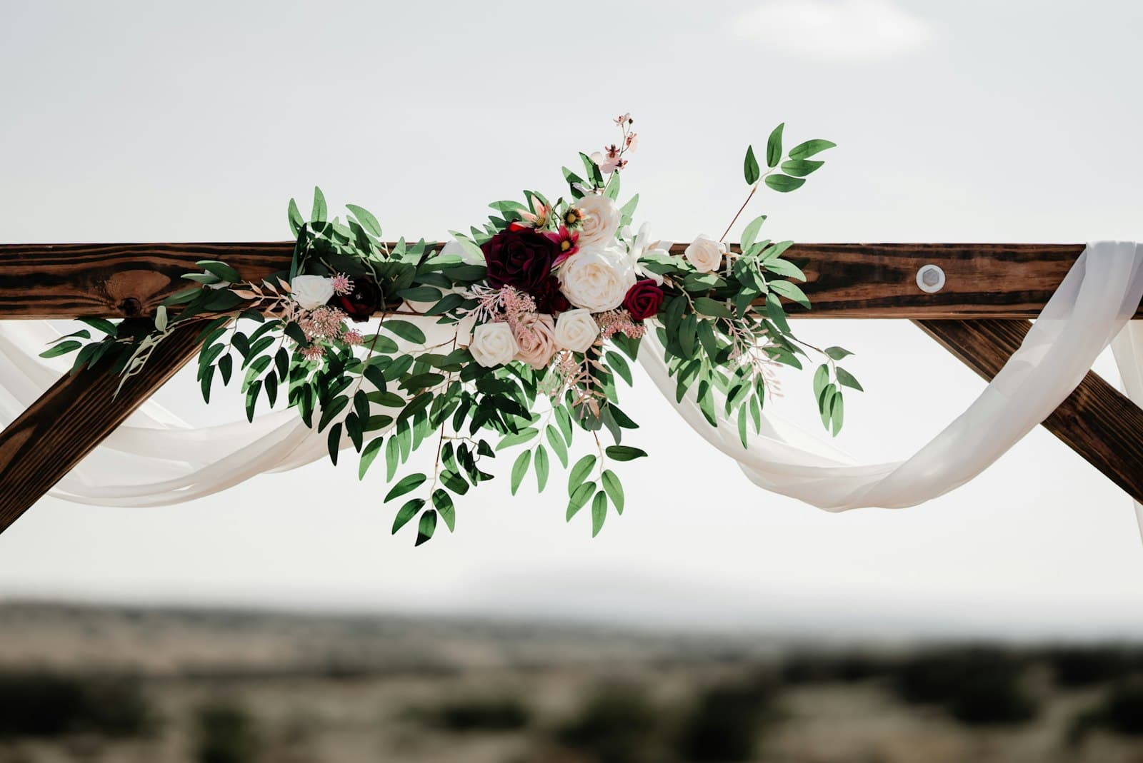 Arche de mariage en bois décorée de roses, de verdure et d’un voilage blanc, devant un paysage